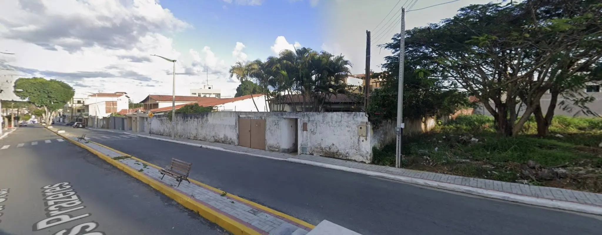 Fachada de uma casa com muro branco, portão de ferro, árvores no interior e terreno baldio no lado esquerdo.