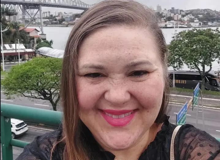 Uma mulher de pele clara com sardas e cabelos castanhos sorri para a selfie, usando um batom rosa vibrante e blusa preta de renda. Ela está em um local elevado, com uma grade verde à esquerda, tendo ao fundo uma ponte metálica sobre um rio e uma via com carros e árvores. A iluminação é natural e o clima parece nublado, destacando a paisagem urbana litorânea atrás dela.