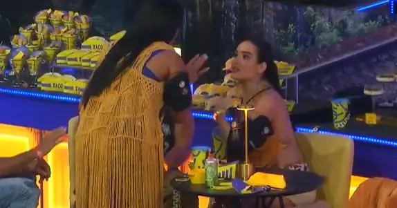 Em uma festa iluminada por luzes amarelas e azuis, duas mulheres conversam intensamente sentadas à mesa; uma delas, de costas, usa um colete mostarda de franjas, enquanto a outra, de frente, gesticula com expressão séria. Ao fundo, balcões exibem diversos lanches embalados em amarelo, compondo um cenário vibrante e movimentado. A mesa redonda à frente contém bebidas e objetos pessoais sob uma luminária moderna e baixa.