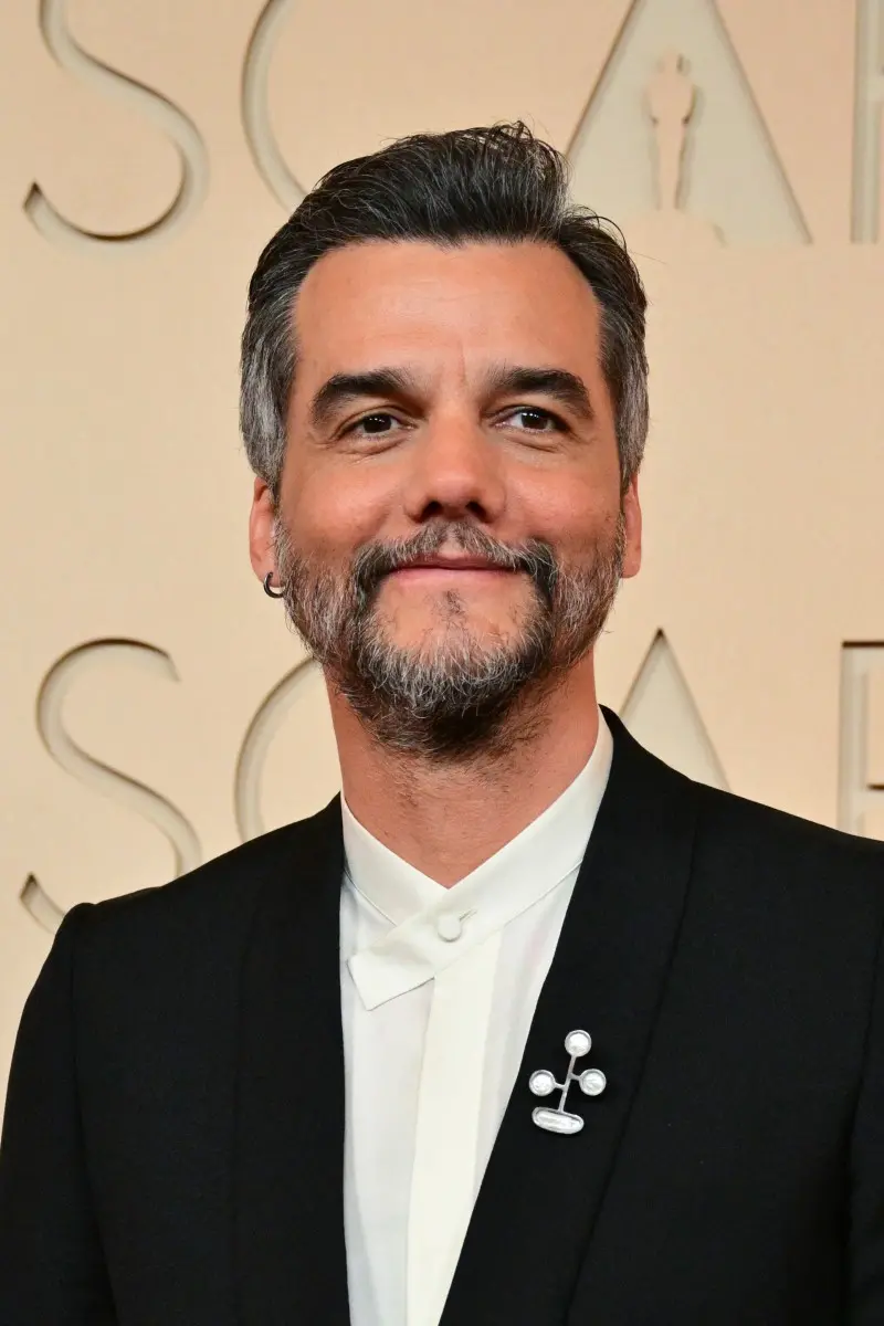 Pessoa vestindo um terno preto com camisa branca de colarinho estruturado, posando diante de um fundo claro com letras grandes em relevo. O terno inclui um broche metálico em formato abstrato, composto por três esferas conectadas por hastes. O enquadramento mostra a parte superior do corpo, com cabelo escuro penteado para cima e presença de um pequeno brinco em uma das orelhas.