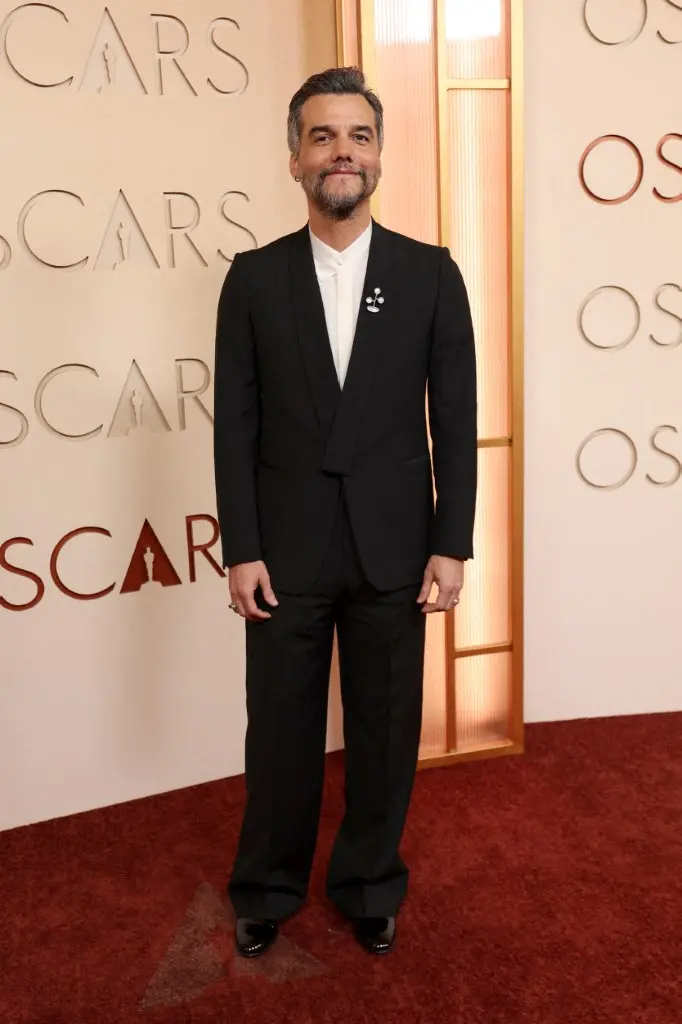 Pessoa em pé sobre um tapete vermelho, diante de um painel claro com a palavra “OSCARS” em grandes letras. A pessoa veste um terno preto de corte clássico com uma camisa branca de gola estruturada. No paletó, há um broche metálico em formato abstrato composto por pequenas esferas conectadas. A iluminação é quente, e ao lado há uma estrutura vertical com painéis translúcidos dourados.