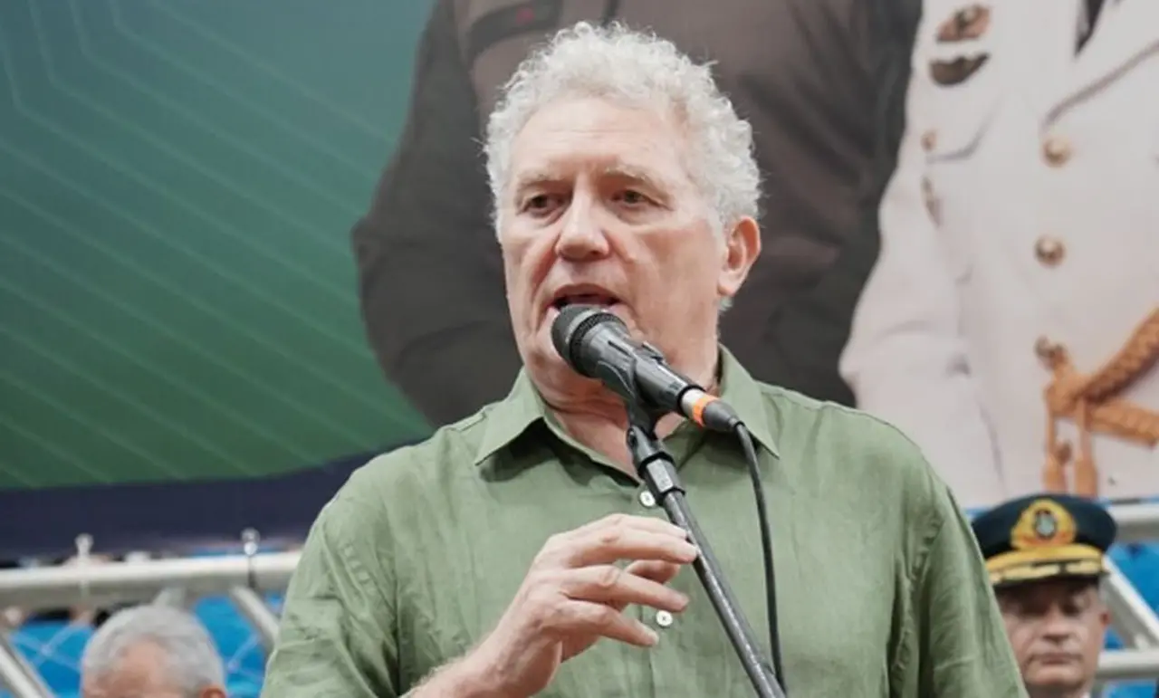 Homem branco de cabelo branco e cacheado, usando blusa de manga na cor verde claro e falando ao microfone.