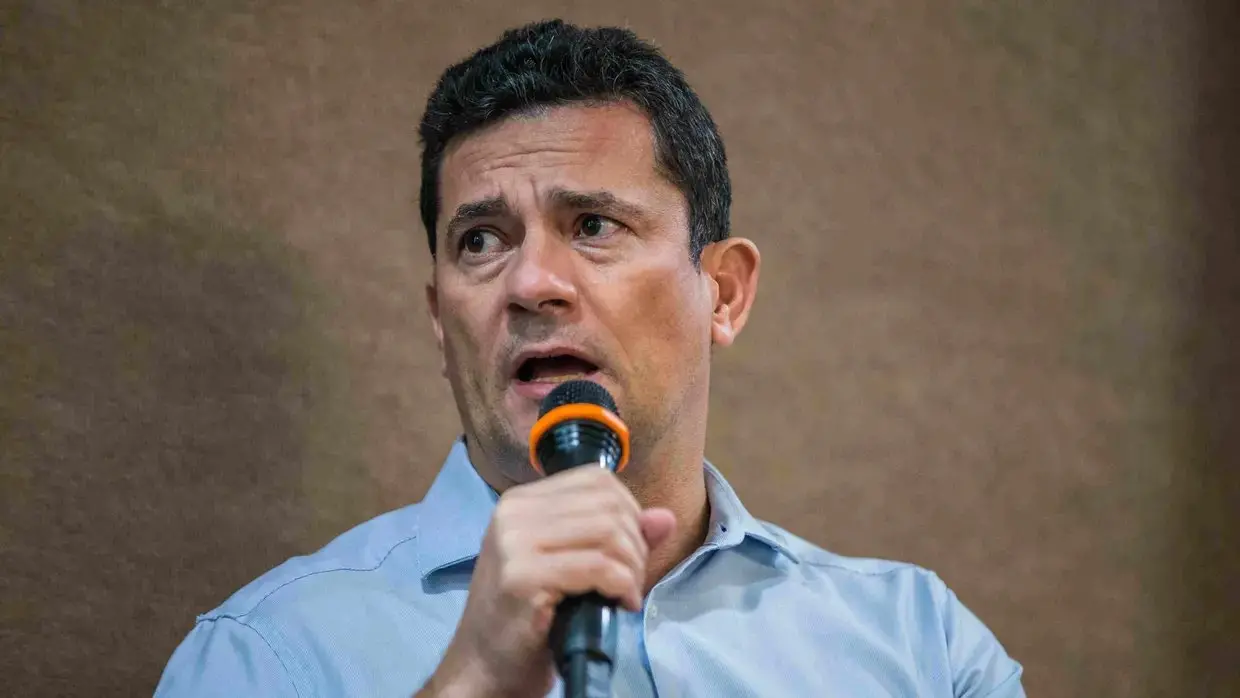 Homem fala ao microfone durante evento, em close, com expressão séria.