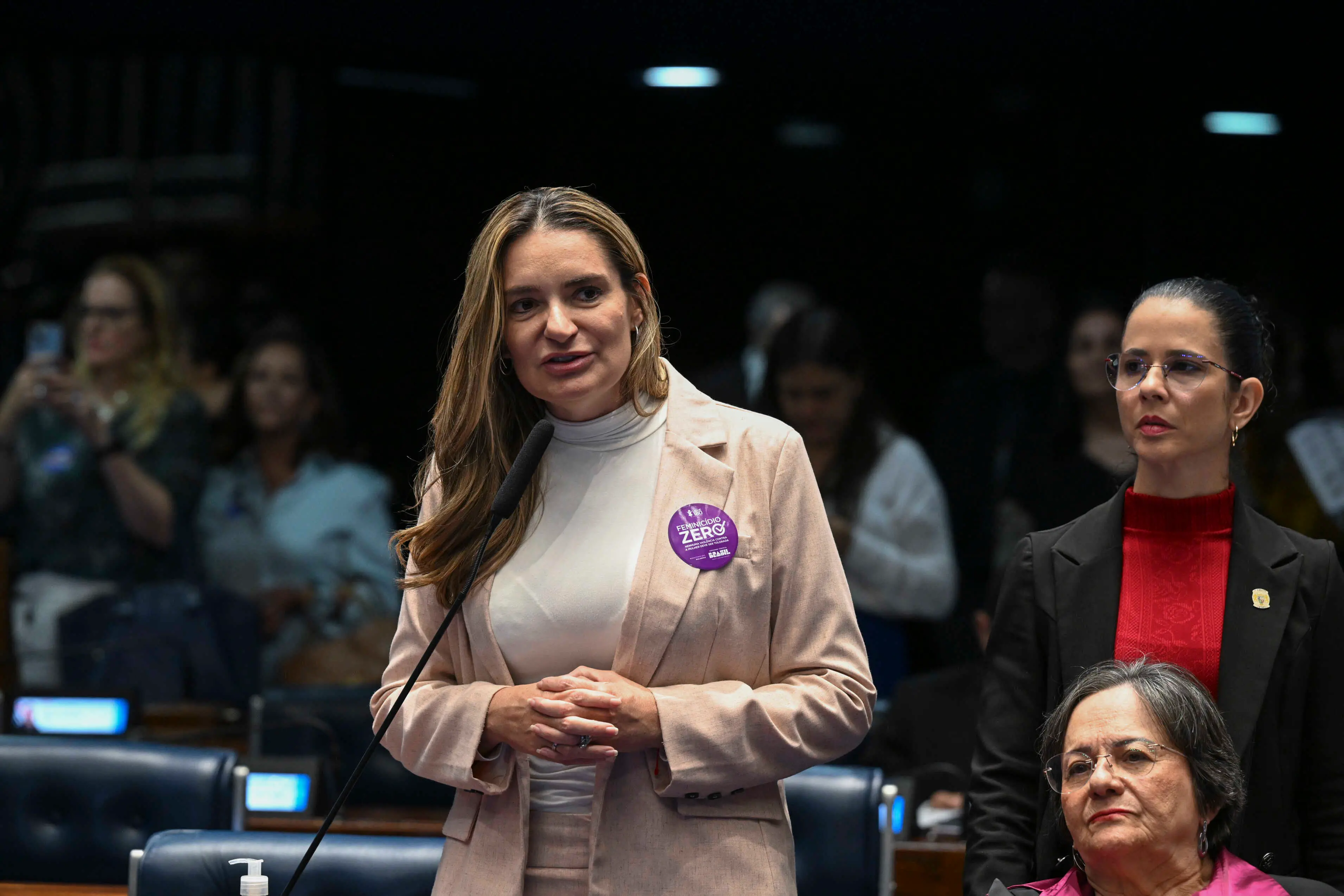 Mulher discursando em um microfone em plenário legislativo, usando blazer bege e um botão roxo escrito ‘Feminicídio Zero’, enquanto duas mulheres estão ao seu lado e outras pessoas aparecem ao fundo.