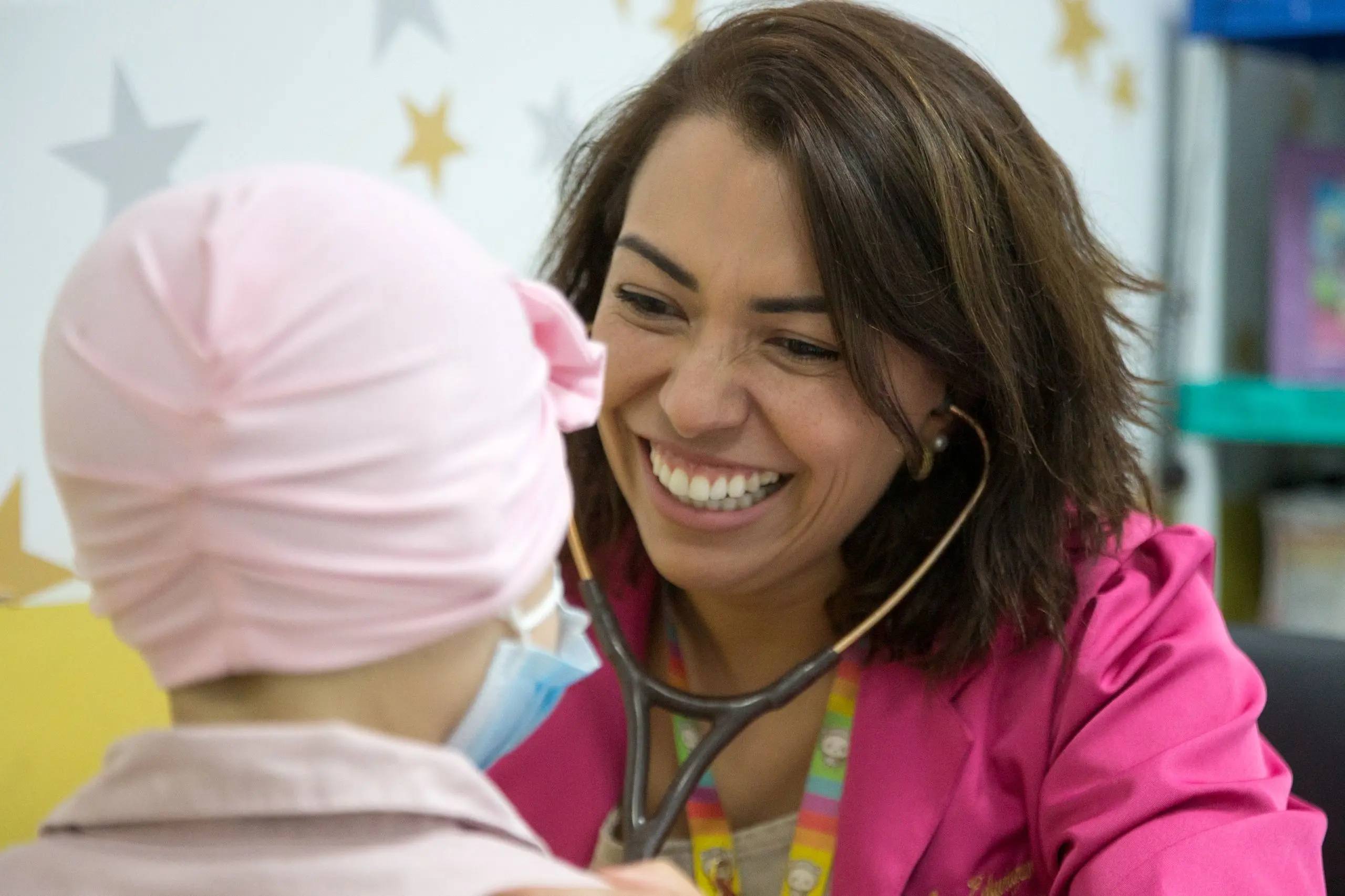 médica Ana Eduvirges Carneiro de Oliveira durante atendimento. ela está sorridente examinando gentilmente uma criança com touca rosa usando um estetoscópio, em um ambiente decorado com estrelas.