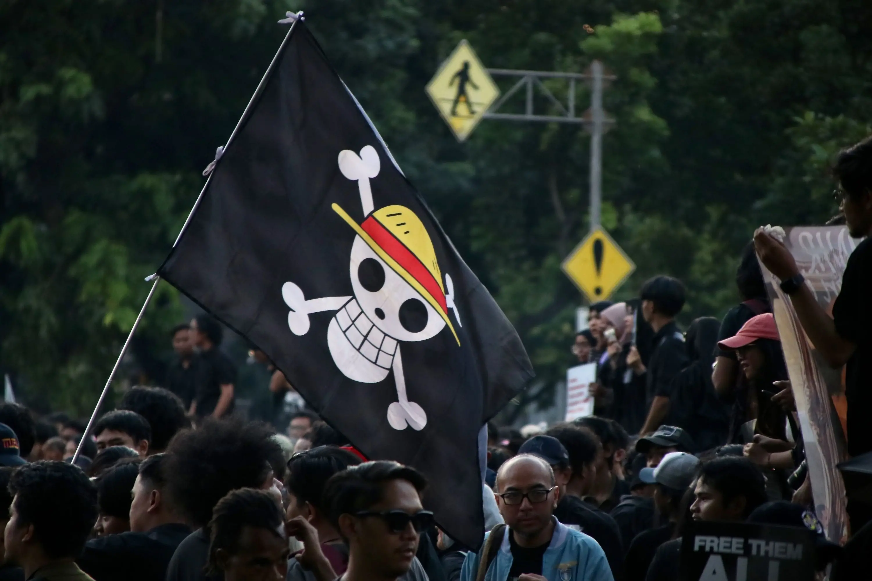 Uma bandeira preta com o símbolo do Jolly Roger dos Piratas do Chapéu de Palha (One Piece) sendo erguida acima de uma multidão de pessoas em um evento ao ar livre.
