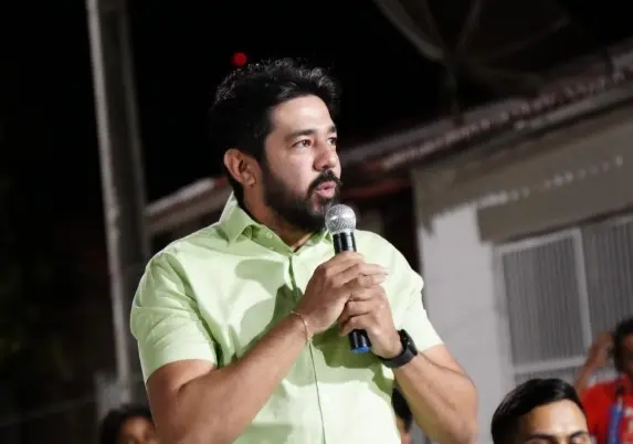 Homem adulto com barba e cabelo escuro fala ao microfone em um evento noturno, usando camisa verde clara e olhando para o lado enquanto discursa.