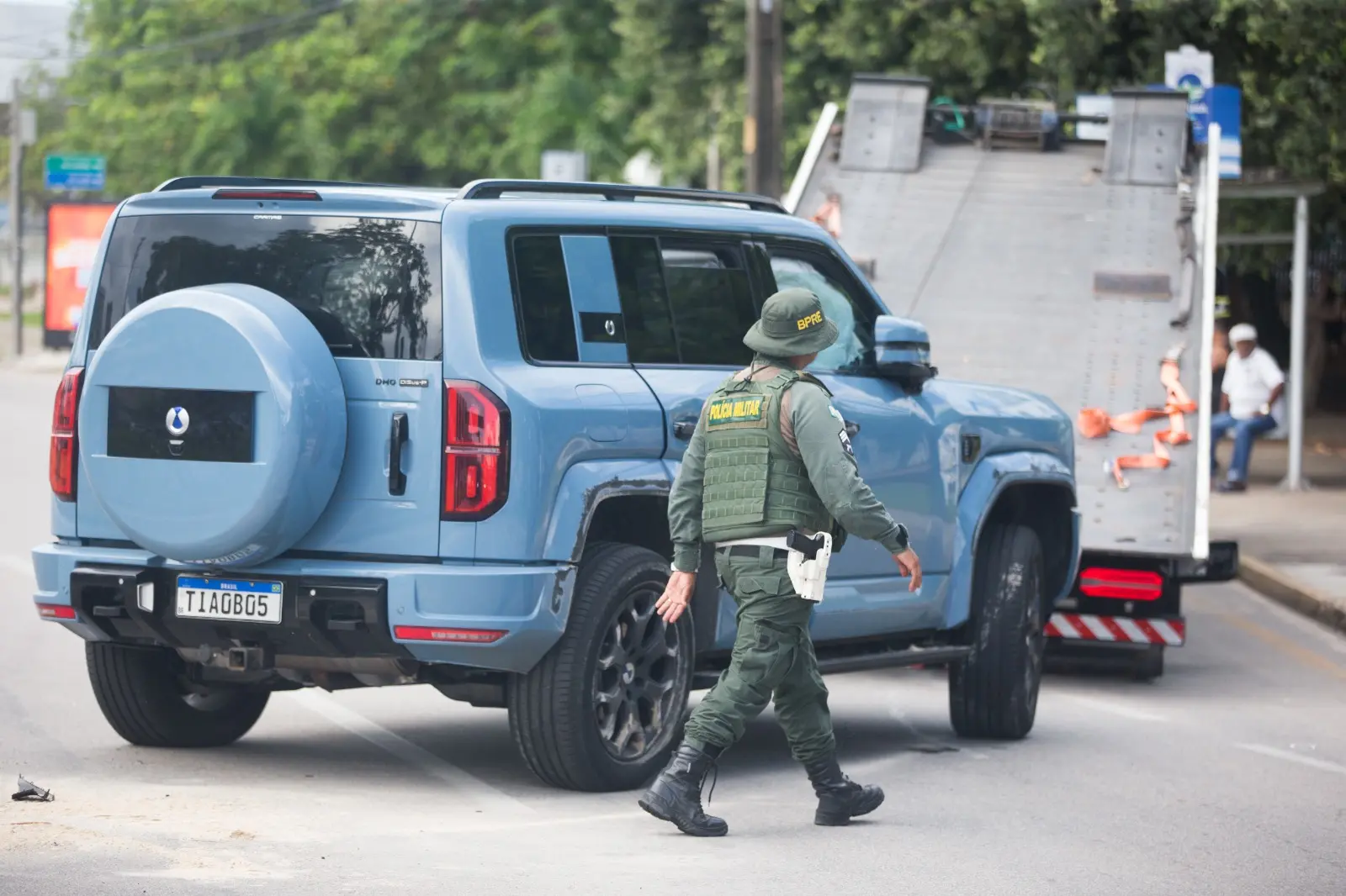 Um policial militar caminha em direção a um SUV azul claro estacionado em frente à rampa de um caminhão guincho em uma via urbana. A cena detalha a fiscalização ou remoção do veículo, com árvores e um ponto de ônibus ao fundo sob a luz do dia.