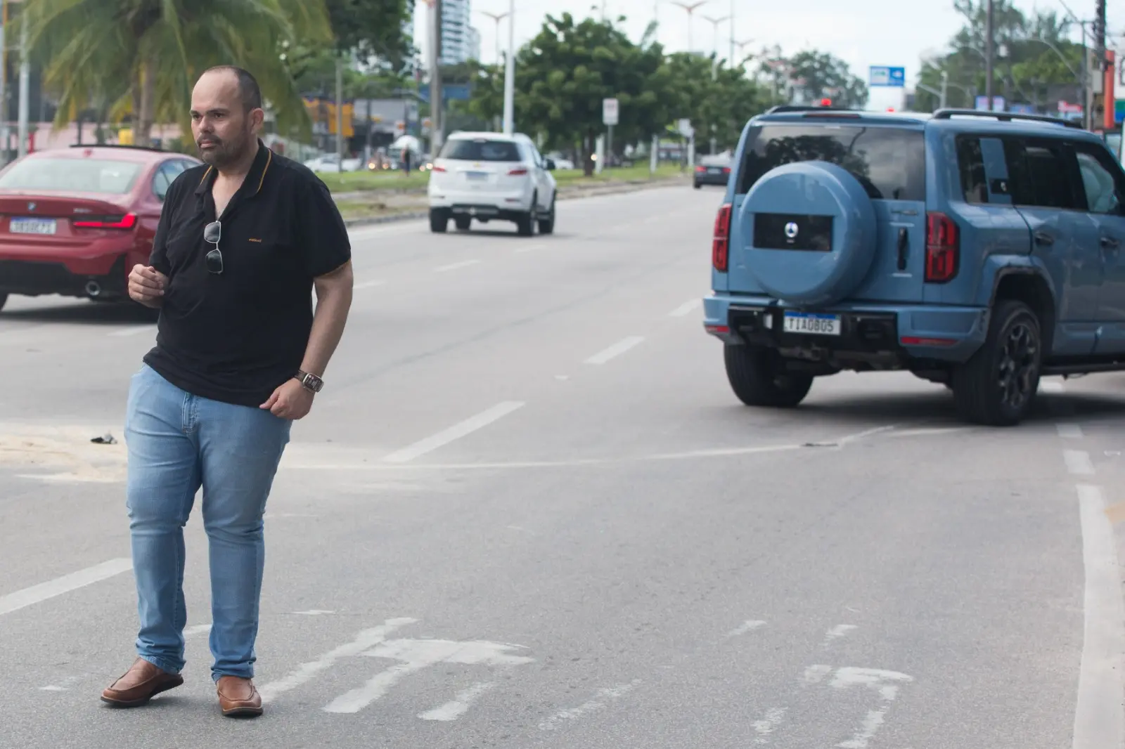 Um homem de camisa polo preta e calça jeans caminha pelo asfalto de uma avenida, com o semblante sério e óculos escuros pendurados no decote. Ao fundo, o SUV azul danificado e outros veículos compõem a cena do acidente sob a luz do dia em um cenário urbano.