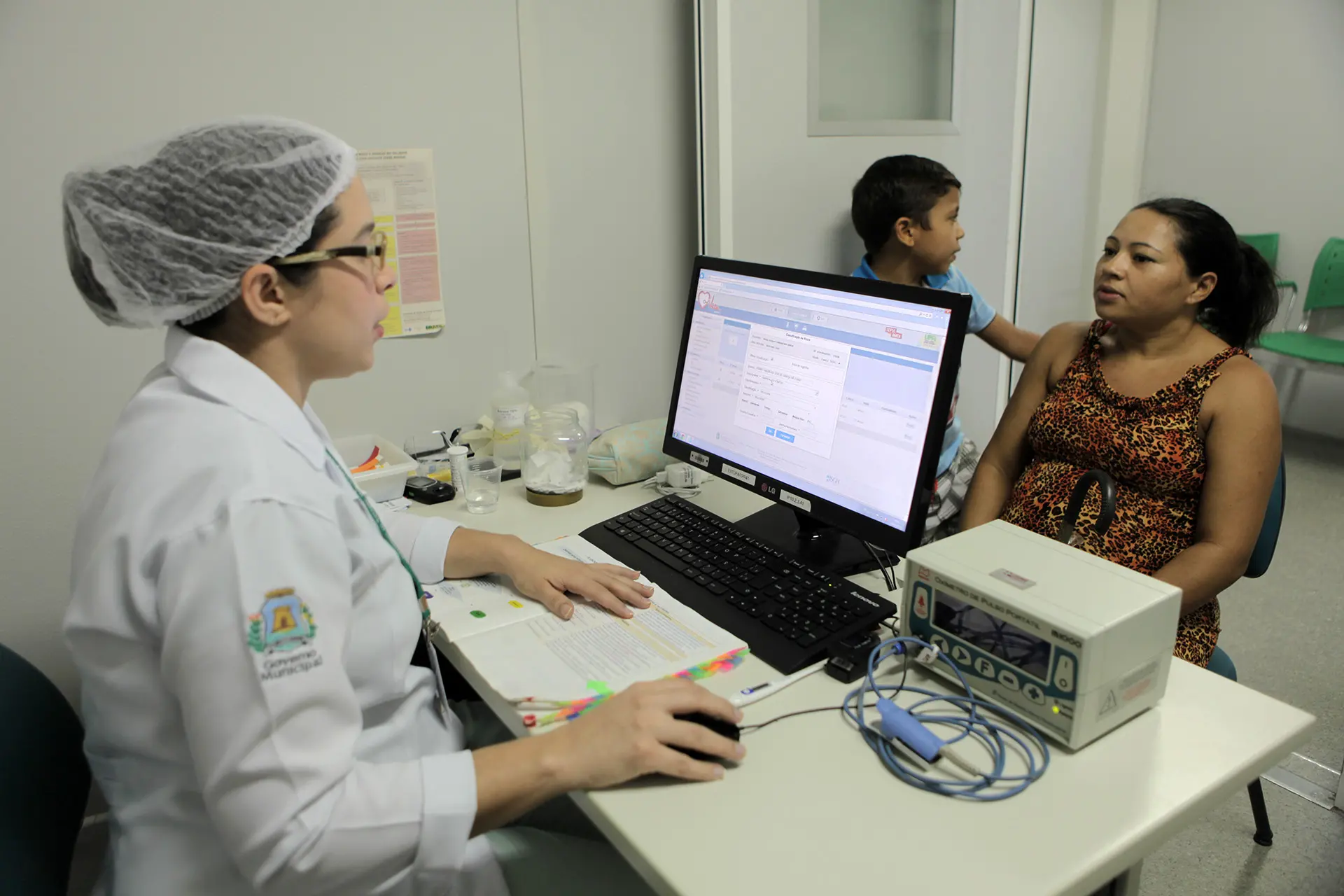 Profissional de saúde realiza atendimento em consultório médico. Ela consulta com paciente e criança em posto de saúde de Fortaleza, representando a saúde primária.