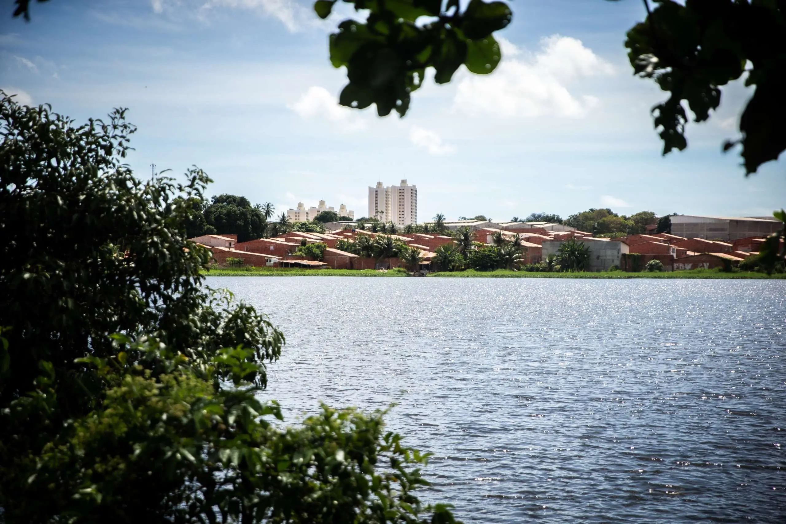 Vista de um açude em Fortaleza com o reflexo do sol, casas de telhado de barro e edifícios altos em segundo plano, emoldurada por folhagem verde.