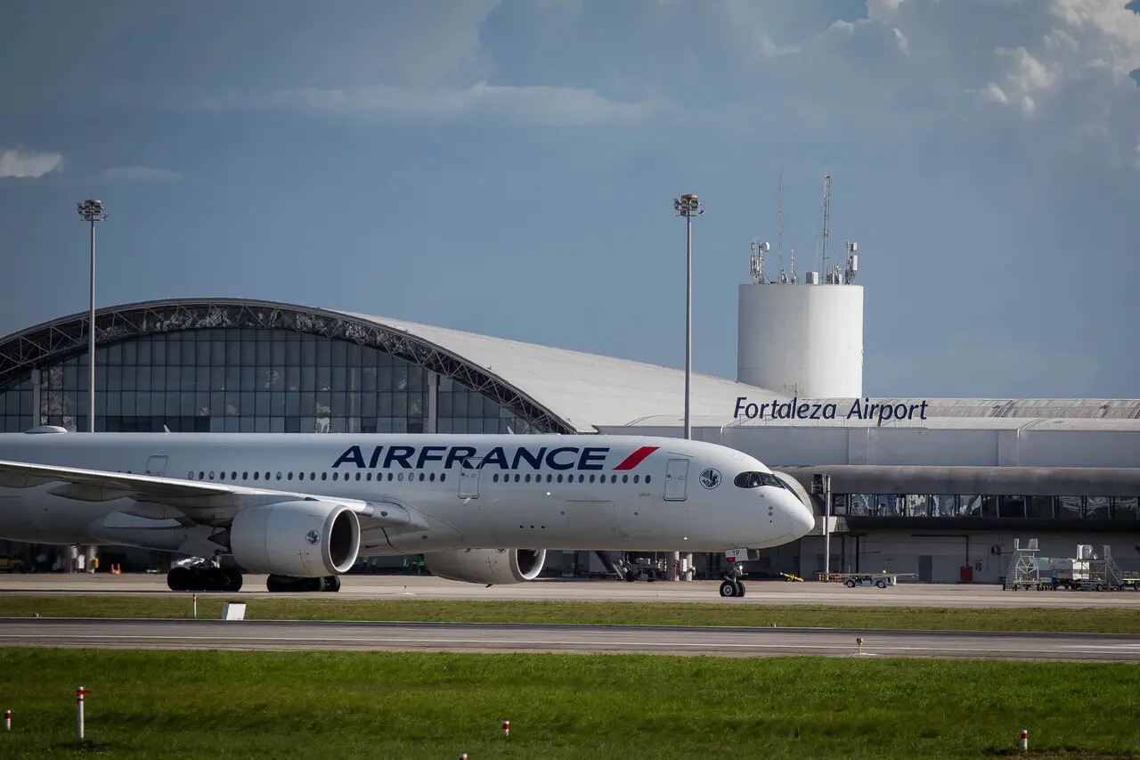 A imagem mostra um avião da Air France estacionado ou taxiando na pista em frente ao terminal do Aeroporto Internacional de Fortaleza - Pinto Martins, em Fortaleza. Ao fundo, aparece o prédio do aeroporto com a inscrição “Fortaleza Airport”, além de torres de comunicação e estruturas do terminal. O avião é um jato comercial de grande porte utilizado em voos internacionais.