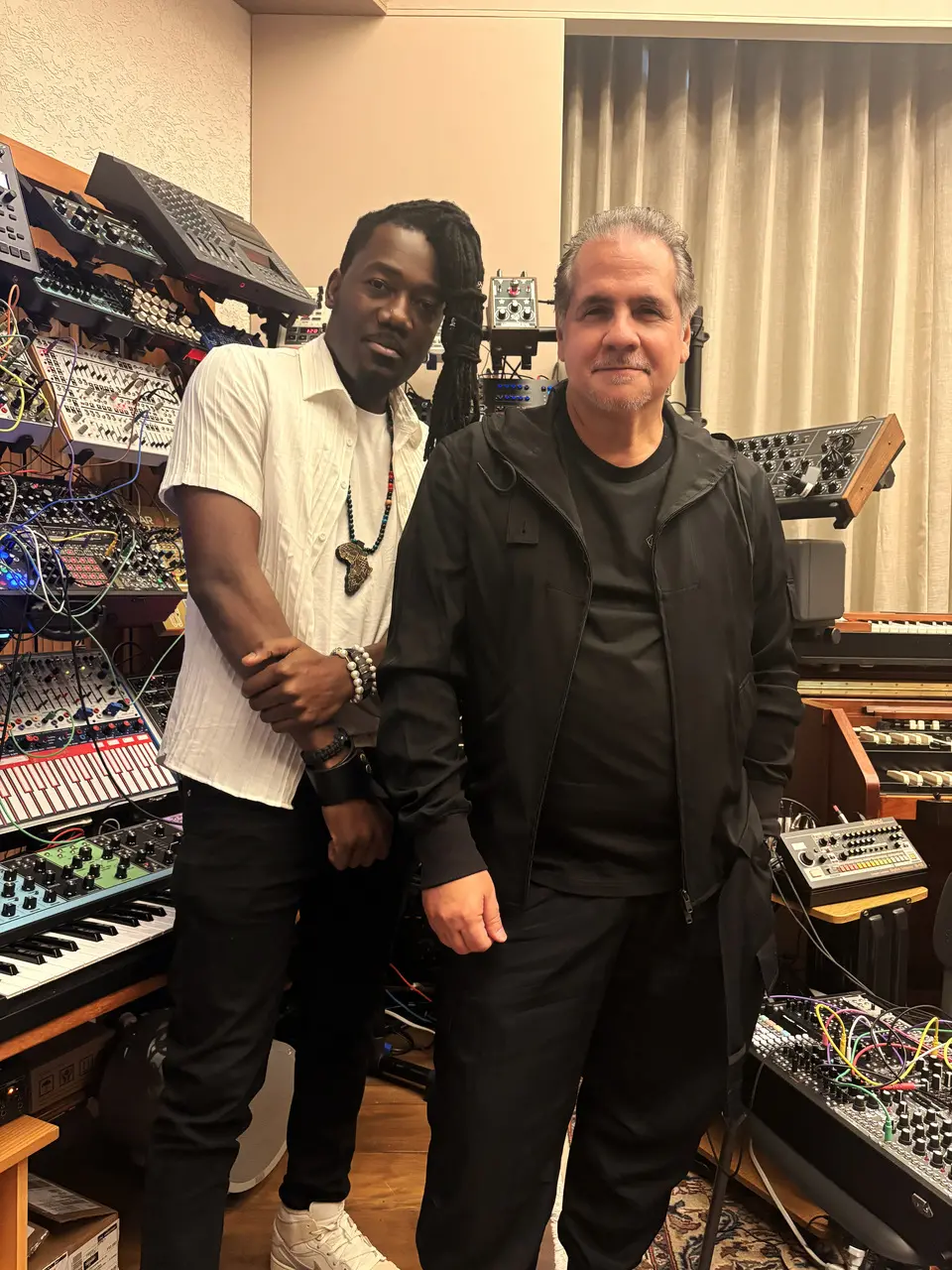 Os músicos Kizaba e Ricardo Bacelar posam lado a lado em um estúdio repleto de sintetizadores modulares e teclados eletrônicos. A imagem destaca a diversidade de equipamentos tecnológicos ao fundo, criando uma atmosfera criativa e profissional que une os dois artistas no centro da composição.