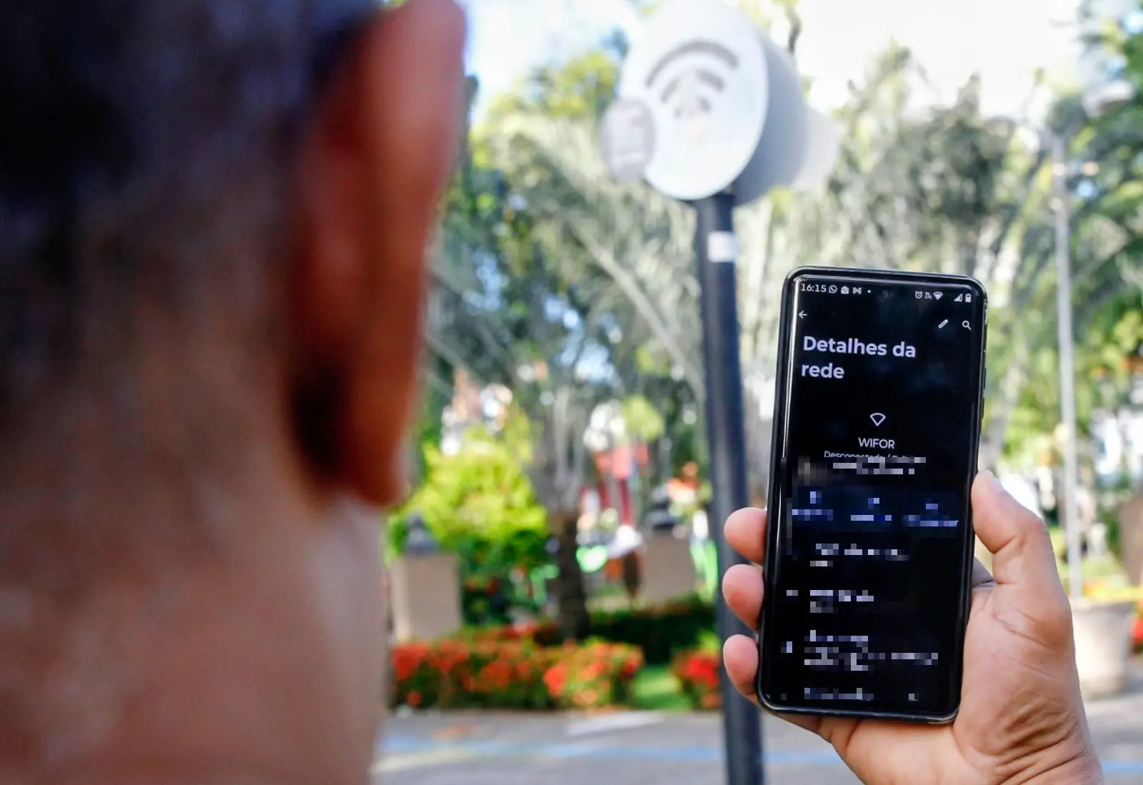 Close-up da parte de trás da cabeça de uma pessoa segurando um smartphone que exibe detalhes da rede, com um poste de Wi-Fi público ao fundo.