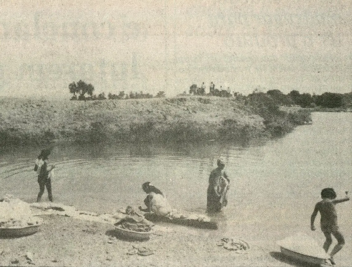 Cena em preto e branco de mulheres e crianças lavando roupas e interagindo na margem do açude Jangurussu, com vegetação e colinas ao fundo.