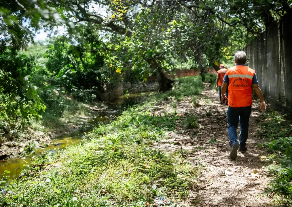 Dois agentes da defesa civil de Fortaleza, com coletes laranjas, caminham por área de vegetação aberta. Ao lado deles, há um canal de água que funciona como sangradouro do açude Jangurussu.