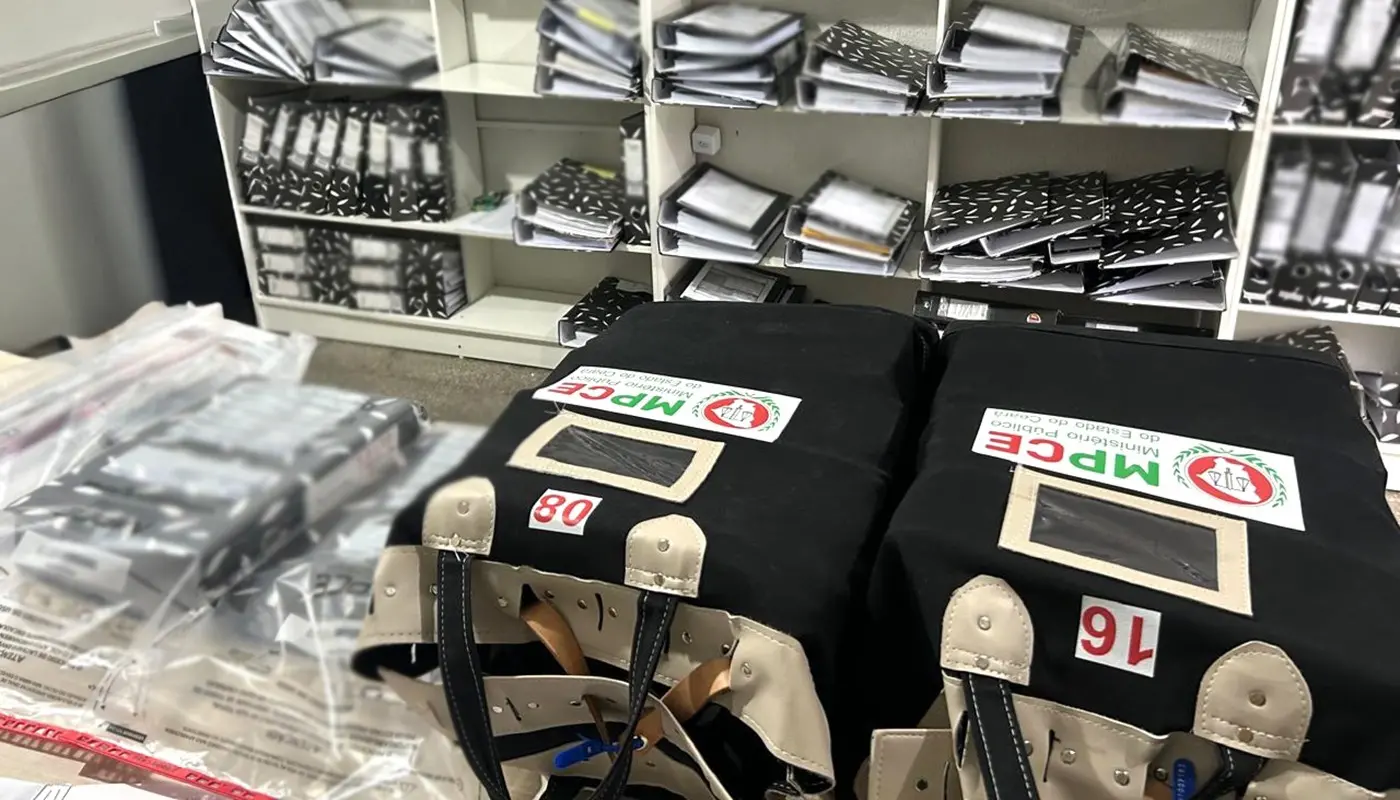Malotes pretos com a logomarca do Ministério Público do Estado do Ceará (MPCE) aparecem em primeiro plano, organizados sobre uma mesa com sacos de evidências. Ao fundo, estantes brancas repletas de pastas de documentos e processos reforçam o cenário de uma investigação oficial em andamento.