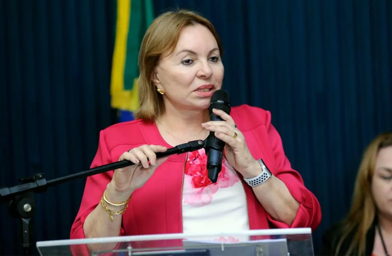 Mulher branca de cabelo liso, curto e loiro, usando blusa social branca e jaqueta rosa, enquanto discursa com um microfone em uma tribun de plenário.