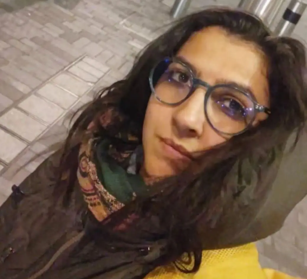 Uma pessoa usando roupas de frio, incluindo um casaco escuro e um cachecol estampado com cores como verde e tons terrosos, aparece em uma área externa pavimentada com blocos retangulares. A pessoa está posicionada próxima ao canto inferior da imagem, e parte de um objeto amarelo aparece em primeiro plano. Ao fundo, há estruturas metálicas verticais iluminadas por luz artificial.