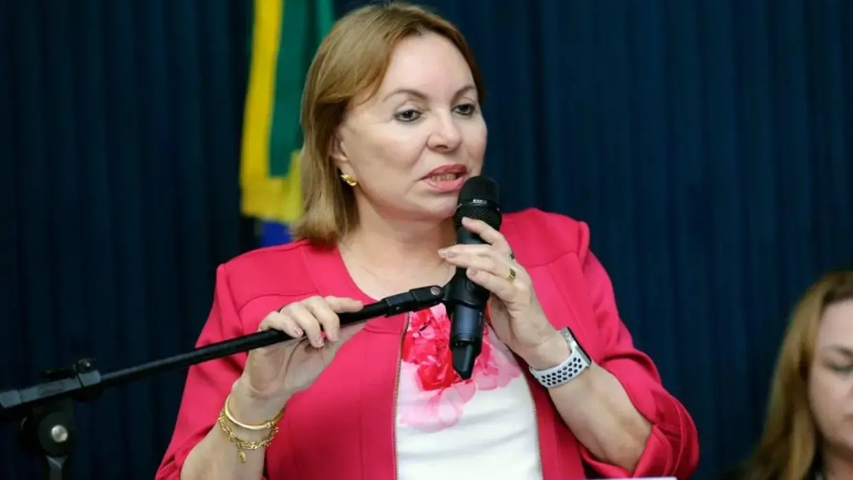Mulher fala ao microfone durante evento, usando blazer rosa, com bandeira do Brasil ao fundo.
