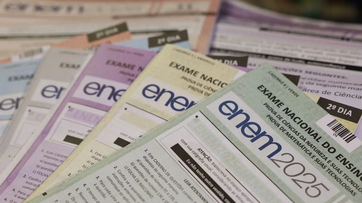 Cadernos de prova do Enem 2025 em diversas cores, como verde, amarelo, rosa e cinza, aparecem organizados e sobrepostos em primeiro plano. O foco destaca o título 