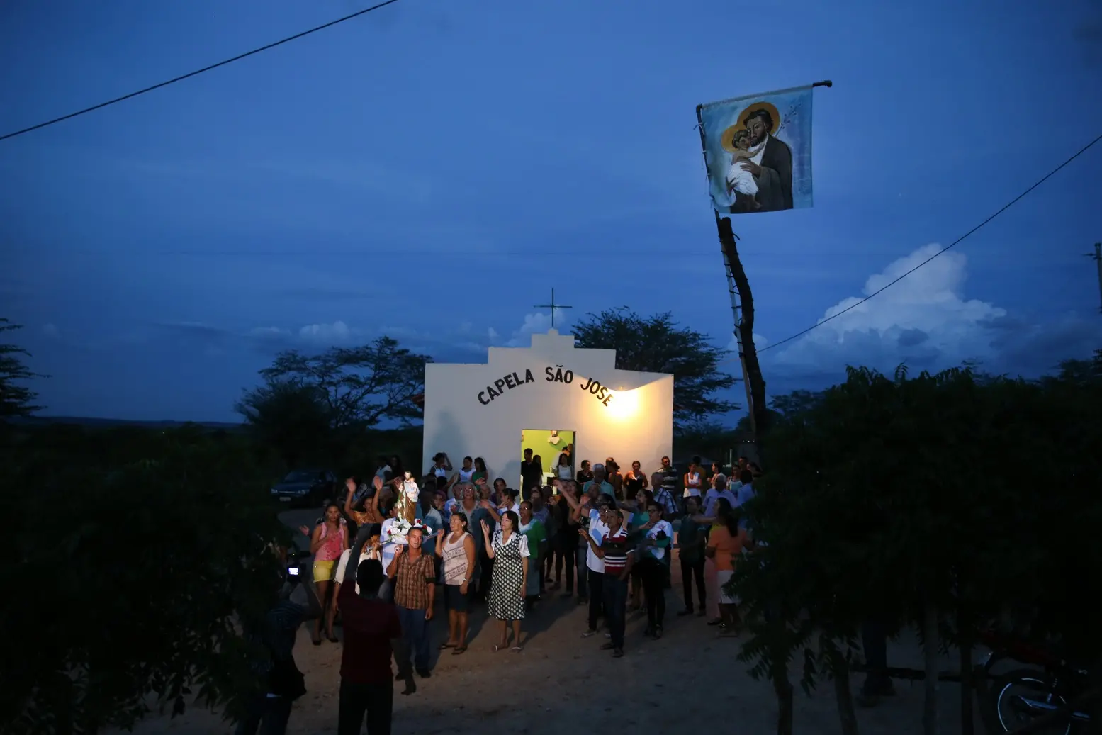Grupo de pessoas reunidas em frente à Capela São José ao anoitecer, com uma bandeira de São José e o Menino Jesus em um mastro.