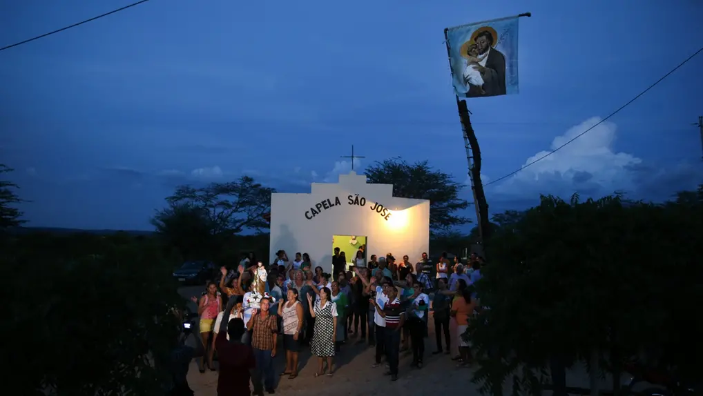 Grupo de pessoas reunidas em frente à Capela São José ao anoitecer, com uma bandeira de São José e o Menino Jesus em um mastro.