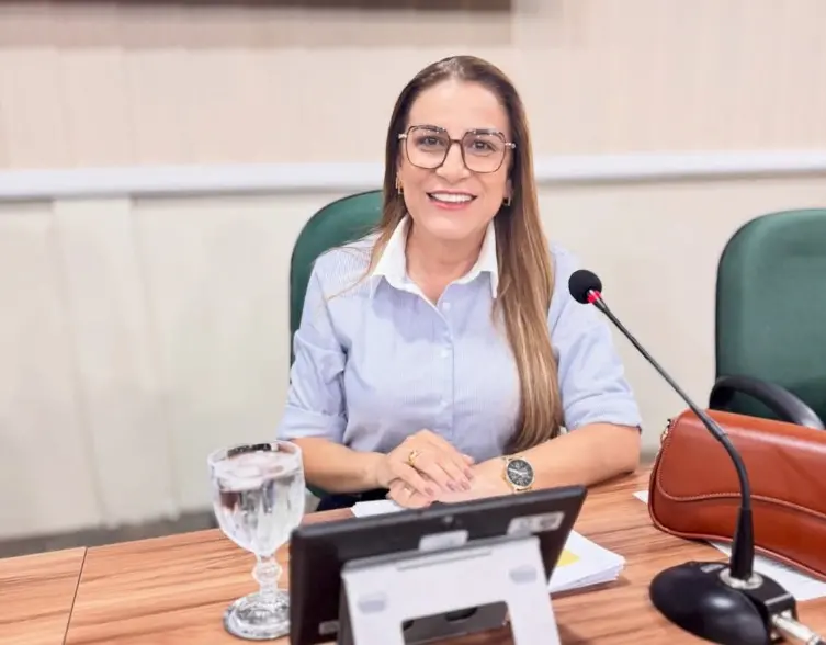 Mulher sorridente, de óculos e cabelos longos lisos, sentada à mesa em um ambiente de reunião. Ela usa camisa social clara e está posicionada diante de um microfone, com um copo de água, um dispositivo eletrônico e uma bolsa sobre a mesa. O fundo mostra cadeiras verdes, sugerindo uma sala institucional ou de conferências.