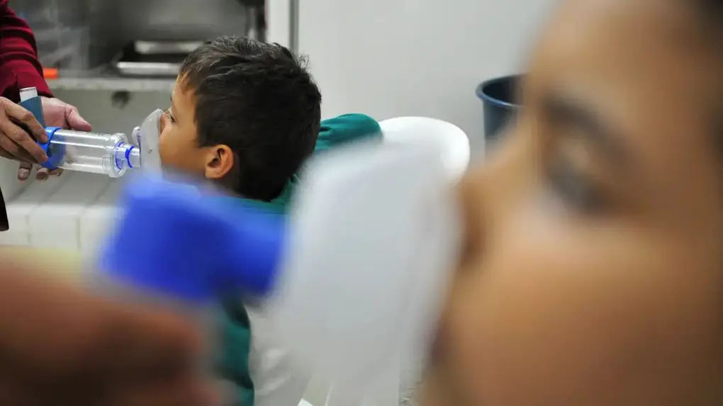 Imagem mostra crianças sentadas recebendo tratamento de nebulização (inalação) com máscara e espaçador em ambiente de saúde. Em primeiro plano, desfocada, outra pessoa segura um inalador, representando o cuidado com doenças respiratórias infantis como influenza ou bronquite.