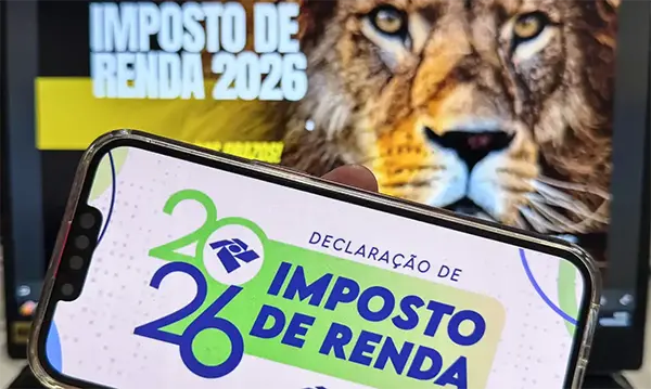 A imagem mostra um celular exibindo a tela da declaração do Imposto de Renda 2026, com o logo “IR 2026” e a frase “Declaração de Imposto de Renda”. Ao fundo, aparece um computador ou TV com a mensagem “Imposto de Renda 2026” e a imagem de um leão — símbolo tradicional da Receita Federal do Brasil.