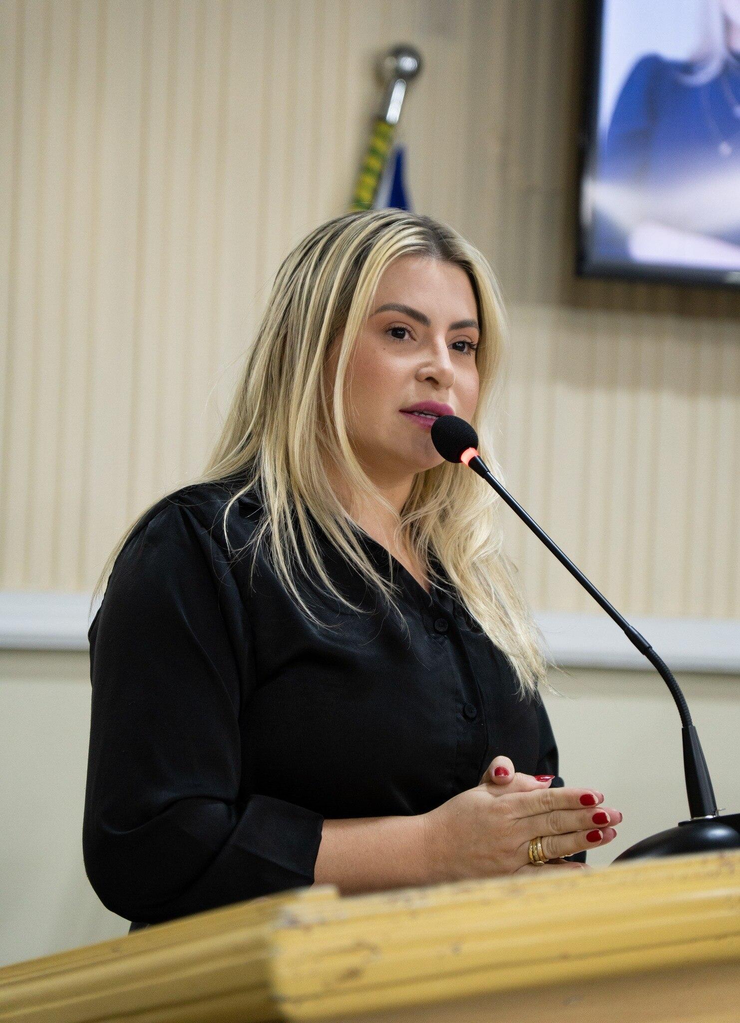 Mulher de cabelo loiro, liso e na altura dos ombros, vestindo blusa preta, fala ao microfone em um púlpito de madeira. Ela está em um ambiente interno que parece uma sala institucional ou plenário, com parede clara ao fundo e uma bandeira parcialmente visível atrás. A mulher mantém expressão séria e concentrada, com as mãos juntas à frente, enquanto se dirige ao público.