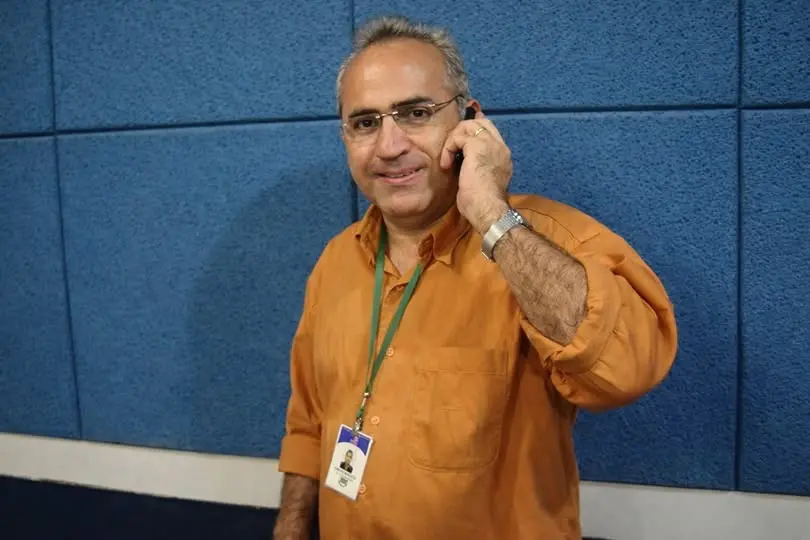 Homem sorridente usando óculos, camisa laranja, com crachá e falando ao telefone, em ambiente de escritório com parede azul ao fundo.