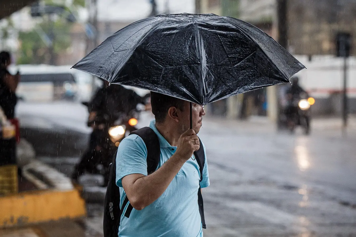 Homem com camisa azul claro e mochila preta segurando um guarda-chuva preto enquanto caminha sob chuva forte em uma rua urbana molhada.