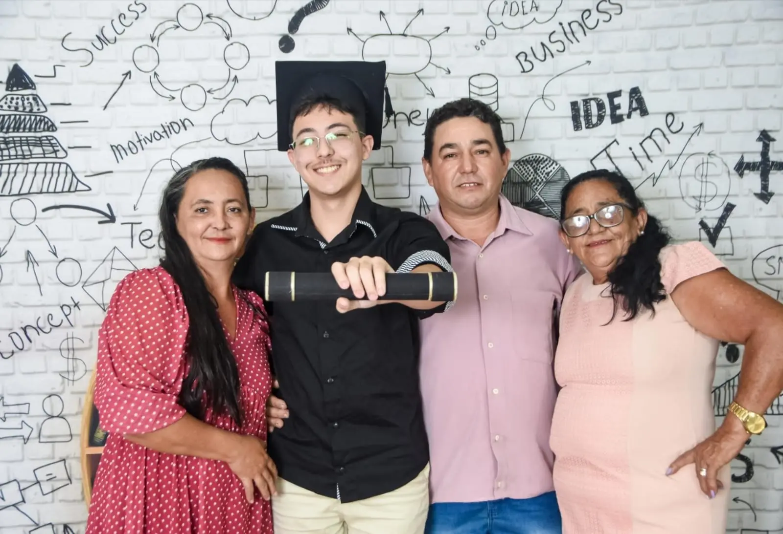 Um jovem sorridente, vestindo beca e capelo, segura seu diploma à frente de três familiares em um registro de celebração de formatura. Eles posam contra uma parede de tijolos brancos decorada com desenhos e palavras ligadas ao sucesso e inovação, como 