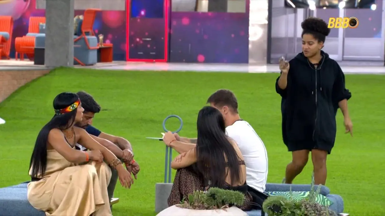 BBB 26: Grupo de particiantes no jardim da cadas. quatro pessoas aparecem sentadas e uma mulher de roupa preta aparece em pé. Milena provocou Cowboy e disse que irá eliminá-lo em um paredão.