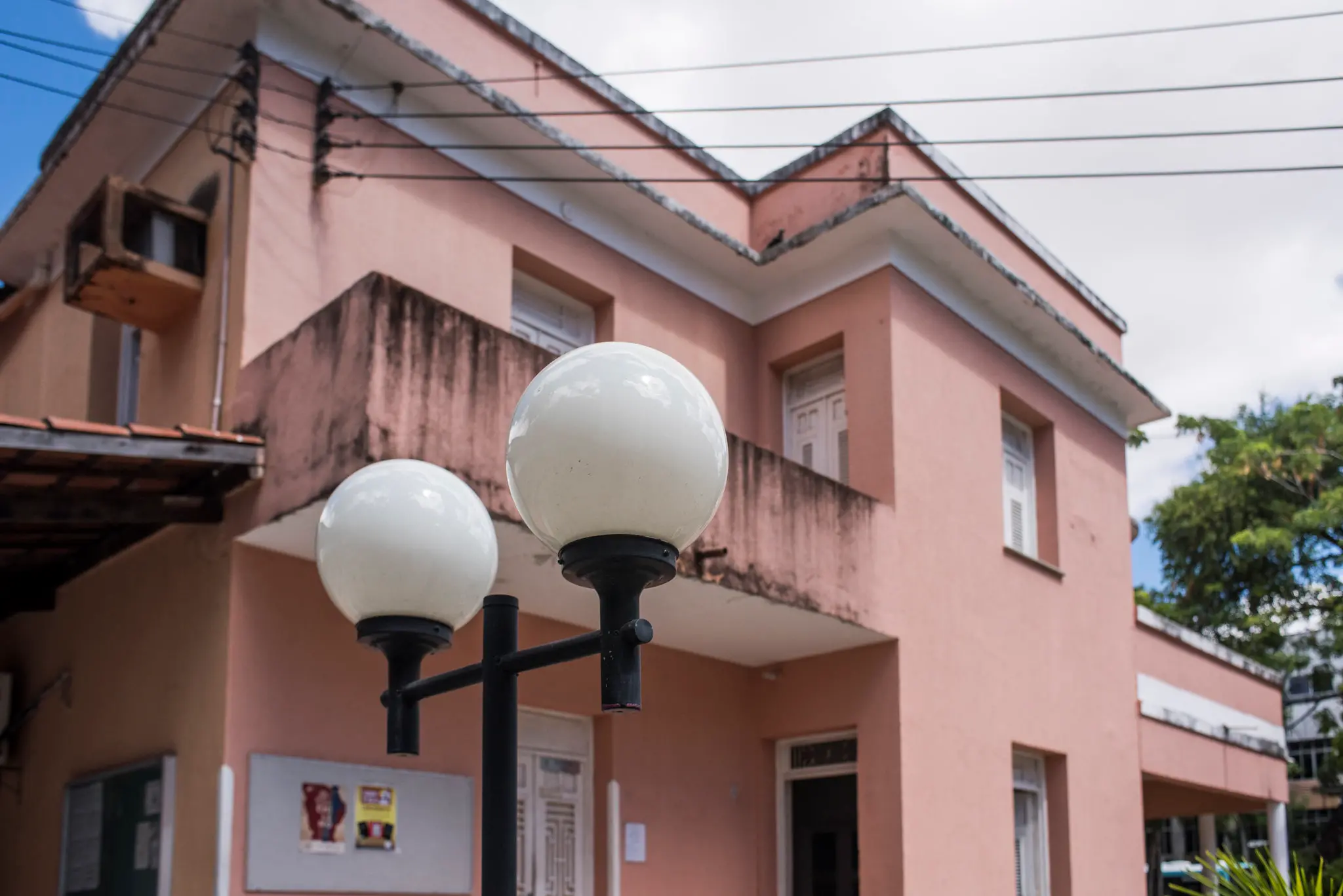 Fachada de um prédio de dois andares, pintado em tom rosado, com áreas escurecidas por umidade nas paredes. Na frente do prédio há um poste de iluminação com três globos brancos. O céu está parcialmente nublado e há árvores ao fundo.