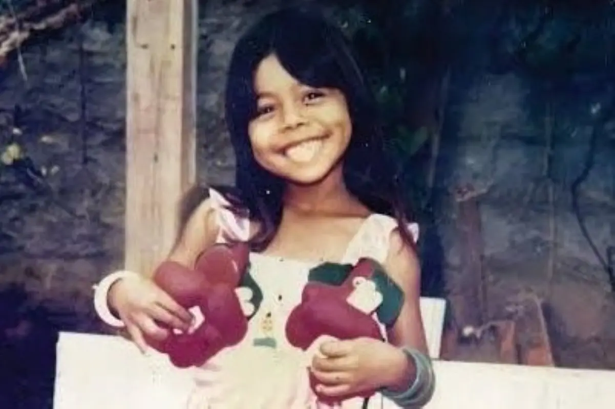 Registro de arquivo pessoal da menina Leide das Neves, uma das vítimas do acidente com césio-137. Na foto, ela sorri alegremente para a câmera, segurando dois grandes objetos decorativos e usa um vestido claro. O registro possui uma estética de fotografia antiga.