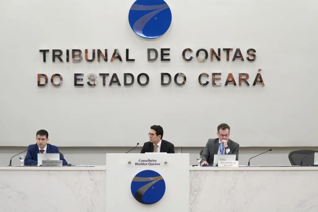 Imagem de uma sala institucional do Tribunal de Contas do Estado do Ceará, com três homens sentados atrás de uma bancada branca durante uma sessão oficial. Ao fundo, na parede, está o nome do órgão em letras grandes. Os participantes usam terno e estão posicionados diante de microfones e documentos, sugerindo uma reunião ou julgamento administrativo.