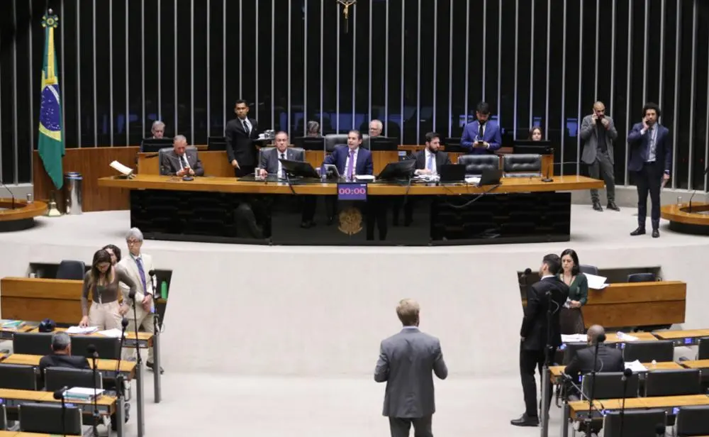 Fotografia do Plenário da Câmara dos Deputados, em Brasília. Em destaque, a mesa diretora ao fundo com parlamentares sentados e um cronômetro digital marcando 00:00. No centro do salão, deputados e assessores circulam entre as bancadas de madeira. À esquerda, vê-se a bandeira do Brasil. A iluminação é clara e o ambiente sugere uma sessão legislativa em andamento.