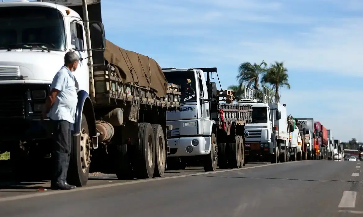 Caminhões enfileirados parados em uma rodovia enquanto um homem de pé observa a cena.