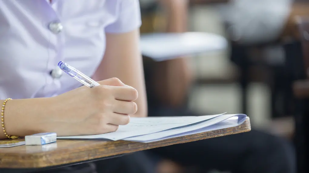 Uma estudante escreve em folhas de prova sobre uma carteira de madeira, segurando uma caneta azul enquanto mantém o foco no exame. O plano fechado destaca suas mãos e o material escolar, com o fundo levemente desfocado revelando o ambiente de uma sala de aula.