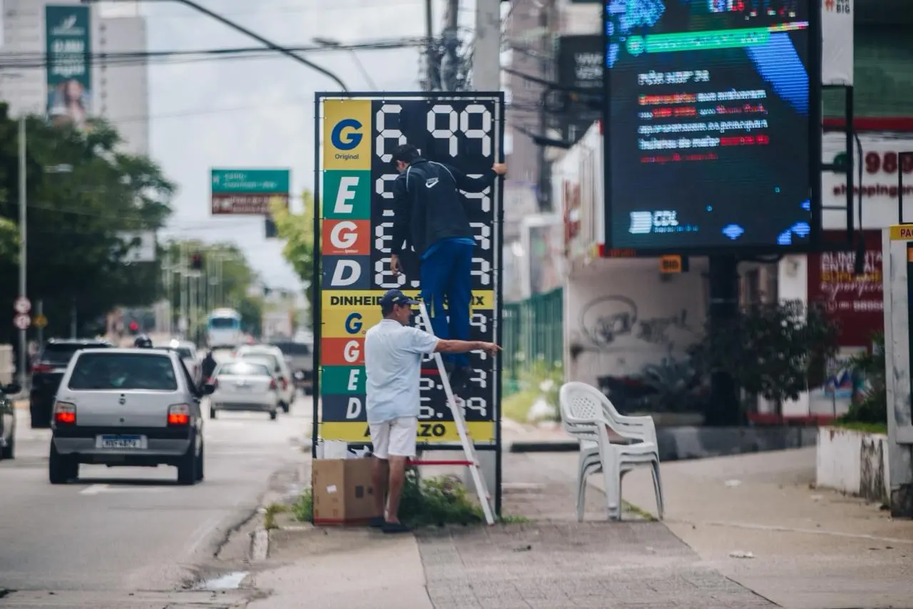 Homens atualizam manualmente placa de preços de combustíveis em avenida movimentada, com carros passando ao lado e painel eletrônico ao fundo.