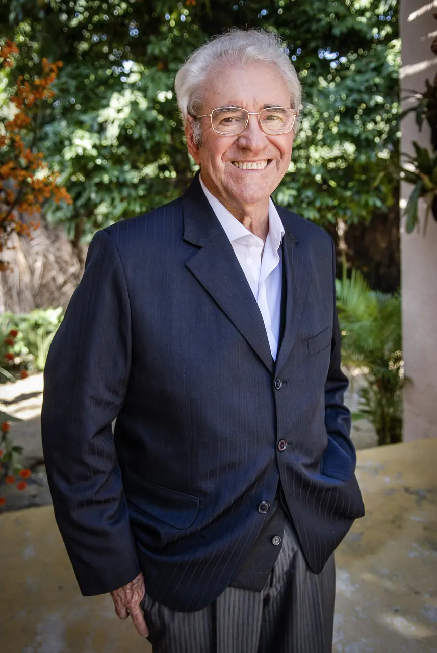 O ator Juca de Oliveira sorri para a câmera vestindo um paletó risca-de-giz escuro sobre uma camisa branca, posando com as mãos nos bolsos. Ao fundo, a vegetação de um jardim iluminado pela luz natural cria um cenário vibrante que complementa a postura elegante do artista.