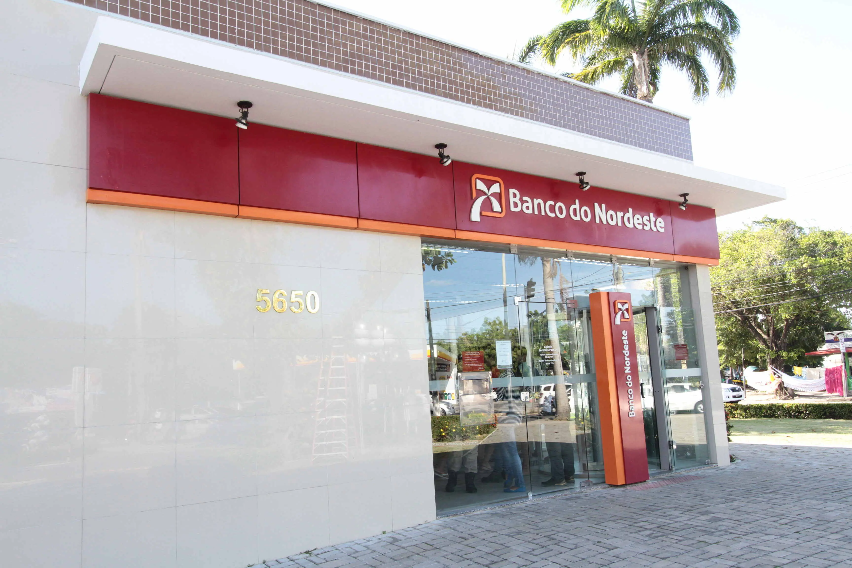 Fachada de uma agência do Banco do Nordeste, com placa vermelha e logotipo acima da entrada. O prédio tem paredes claras, porta de vidro e o número 5650 fixado na parede. Em frente, há calçada de piso intertravado e, ao lado, árvores e uma palmeira visíveis ao fundo.