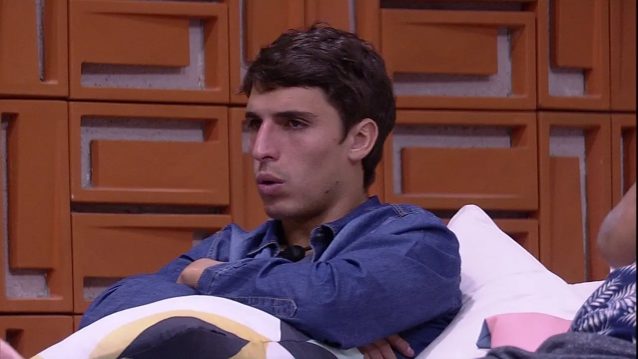 Felipe Prior aparece sentado em um sofá com os braços cruzados e uma expressão de atenção, vestindo uma camisa jeans azul durante o confinamento no Big Brother Brasil. Ao fundo, a parede de cobogós laranjas compõe o cenário da casa.