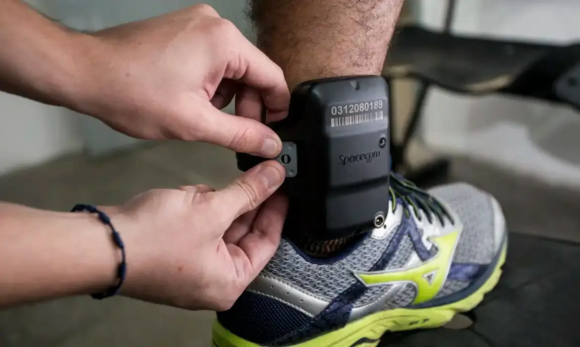 Close-up de uma pessoa ajustando uma tornozeleira eletrônica preta no tornozelo de outra pessoa, que usa tênis esportivo cinza com detalhes verdes, em ambiente interno.