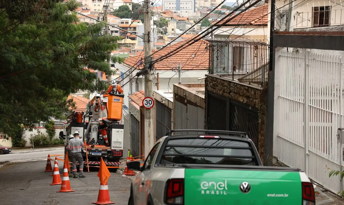 Rua residencial em área urbana com casas de dois andares e telhados de cerâmica. À direita, uma caminhonete da Enel Brasil está estacionada, enquanto, ao fundo, dois trabalhadores uniformizados realizam manutenção em fios elétricos usando um caminhão com cesto elevatório. Cones de sinalização isolam parte da via, e há um poste com limite de velocidade de 30 km/h. Fios e cabos cruzam a cena acima da rua, com árvores e construções ao redor.