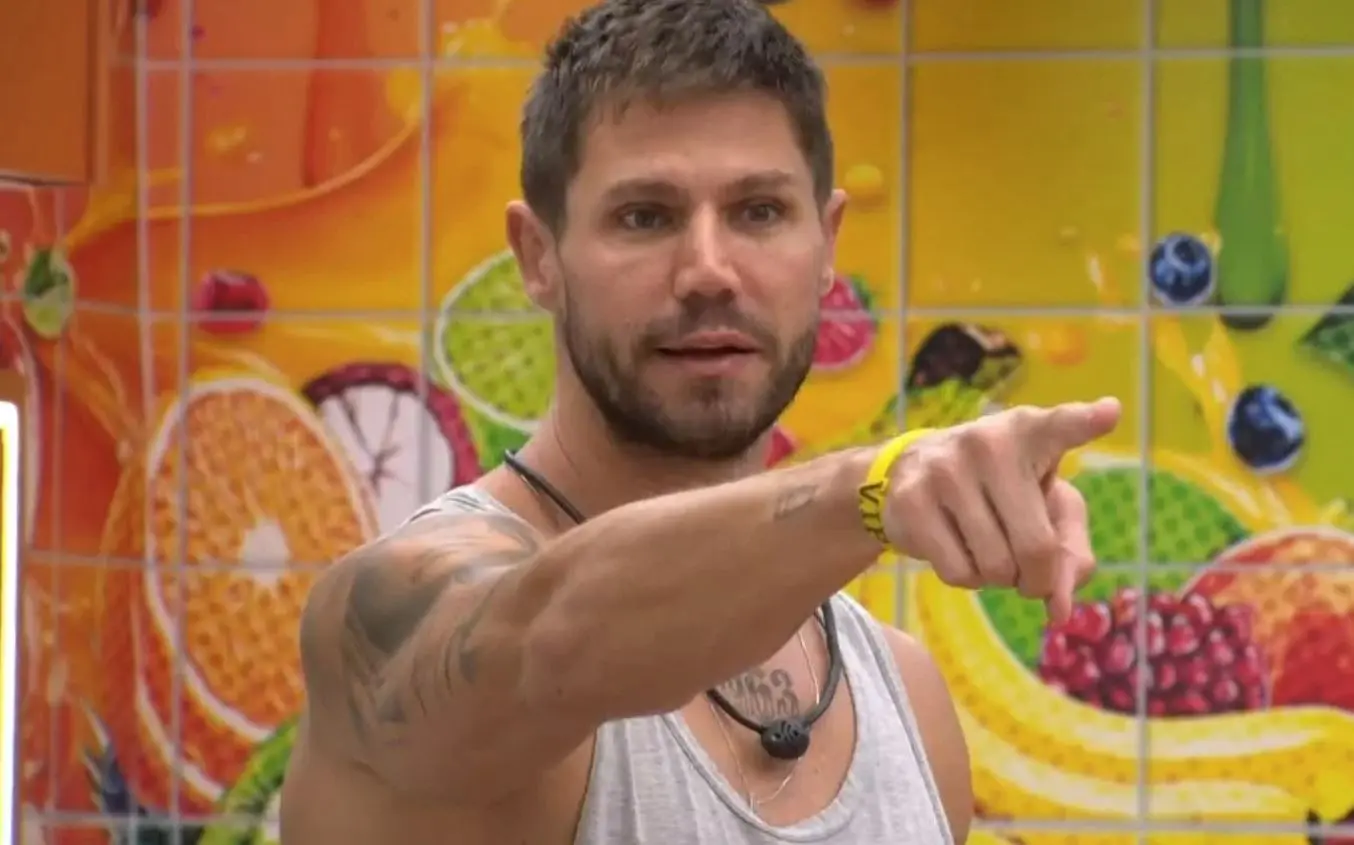 Uma pessoa usando camiseta regata clara está em pé diante de uma parede colorida com ilustrações grandes de frutas. A pessoa aponta com o braço estendido para a direita da imagem, exibindo tatuagens no braço e usando uma pulseira amarela. Um microfone de lapela está preso à roupa.