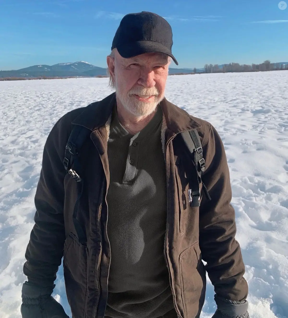 Chuck Norris sorri para a câmera vestindo jaqueta marrom, boné preto e luvas em meio a uma vasta planície coberta de neve. Ao fundo, montanhas distantes e um céu azul limpo completam a paisagem gélida e iluminada pelo sol.