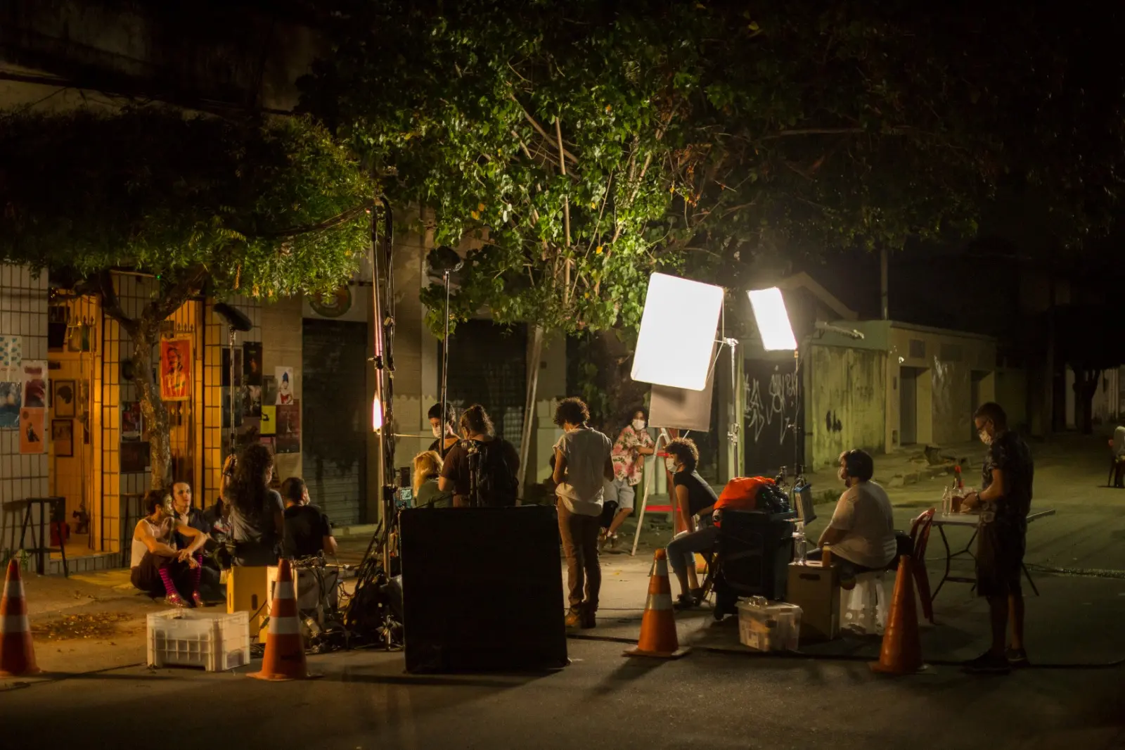 Uma equipe de cinema trabalha em uma gravação noturna do filme cearense 'A Filha do Palhaço' sob a luz de refletores em uma rua de Fortaleza, com atores sentados na calçada ao fundo. A cena captura o contraste entre a escuridão da noite e a iluminação artificial do set, cercado por cones e equipamentos de produção.